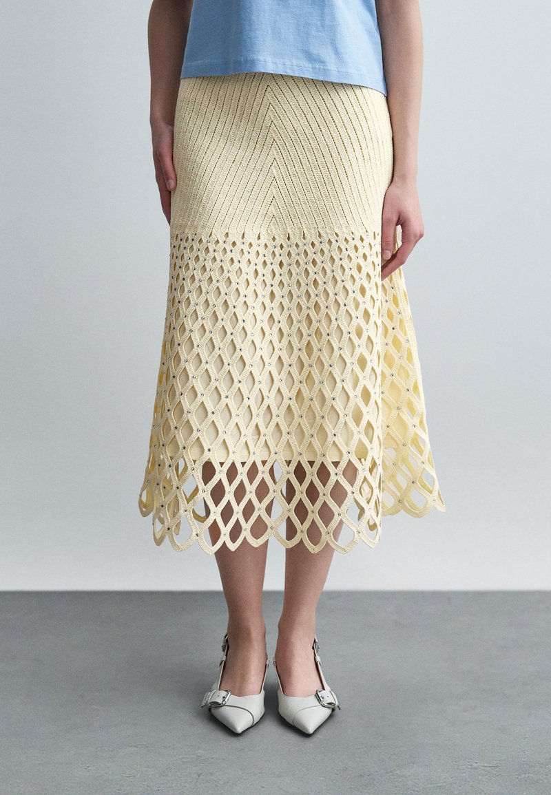 Femme portant une jupe midi tricotée jaune pâle avec un motif ajouré en forme de diamant et des chaussures blanches à boucle pointue, debout sur un sol gris.