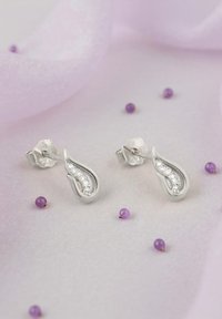 Silver droppformade örhängen med en rad klara stenar på framsidan, mot en mjuk lavendelfärgad bakgrund med små lila kulor.