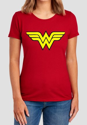 Frau trägt ein rotes T-Shirt mit einem großen gelben Wonder Woman-Logo auf der Brust und blaue Jeans vor einem einfarbigen Hintergrund.