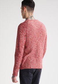 Rödmelerad tröja med rund halsringning, långa ärmar, strukturerat mönster och en liten logotagg vid fållen. Tyget verkar mjukt och avslappnat.