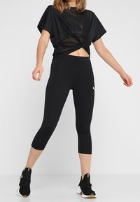 Černý sportovní top se uzlovým detailem, kombinovaný s černými capri legínami a černými vysokými teniskami se zlatými akcenty. Hladká textilie.