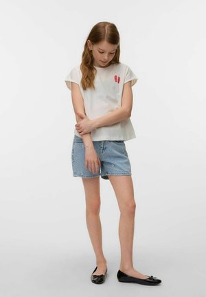 Young girl in rolled-sleeve white shirt with red prints, denim shorts, and black ballet flats standing with arms crossed, looking down.