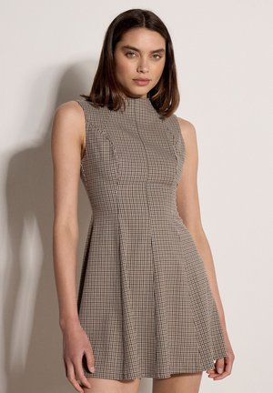 Jeune femme aux cheveux bruns mi-longs portant une robe ajustée sans manches à carreaux, debout devant un fond clair uni.