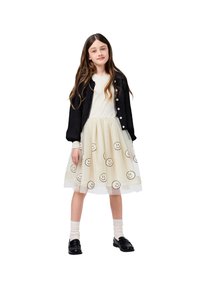 Črn kardigan preko bele majice, slojevita krila z vezenimi nasmejanimi obrazi na tilu, v kombinaciji s črnimi čevlji in belimi nogavicami.