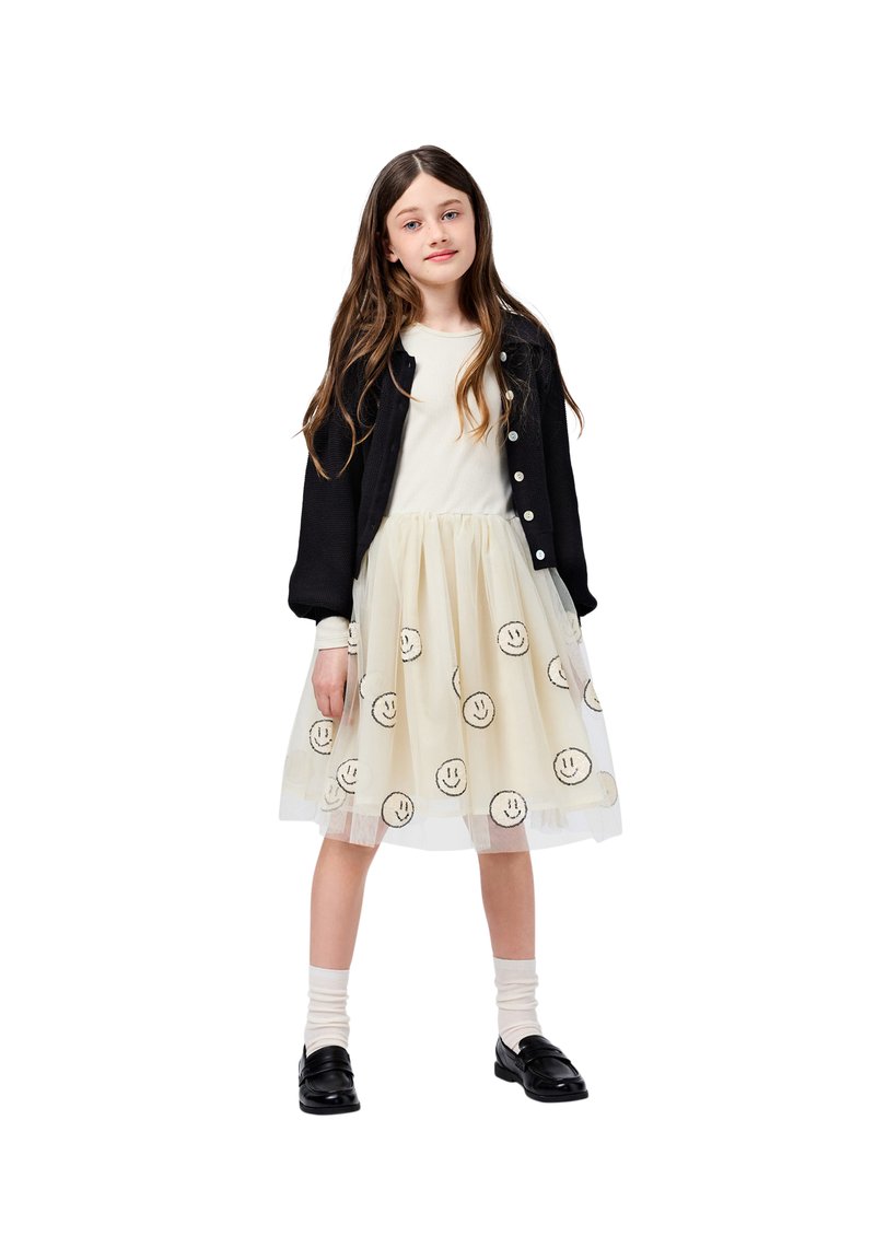 Črn kardigan preko bele majice, slojevita krila z vezenimi nasmejanimi obrazi na tilu, v kombinaciji s črnimi čevlji in belimi nogavicami.