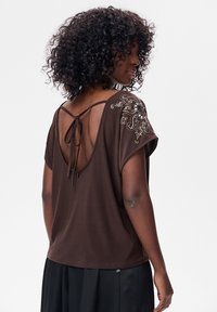 Brązowa bluzka z krótkim rękawem, otwartym tyłem, wiązaniem i ozdobnymi koralikami na ramionach. Miękki materiał i luźny krój.