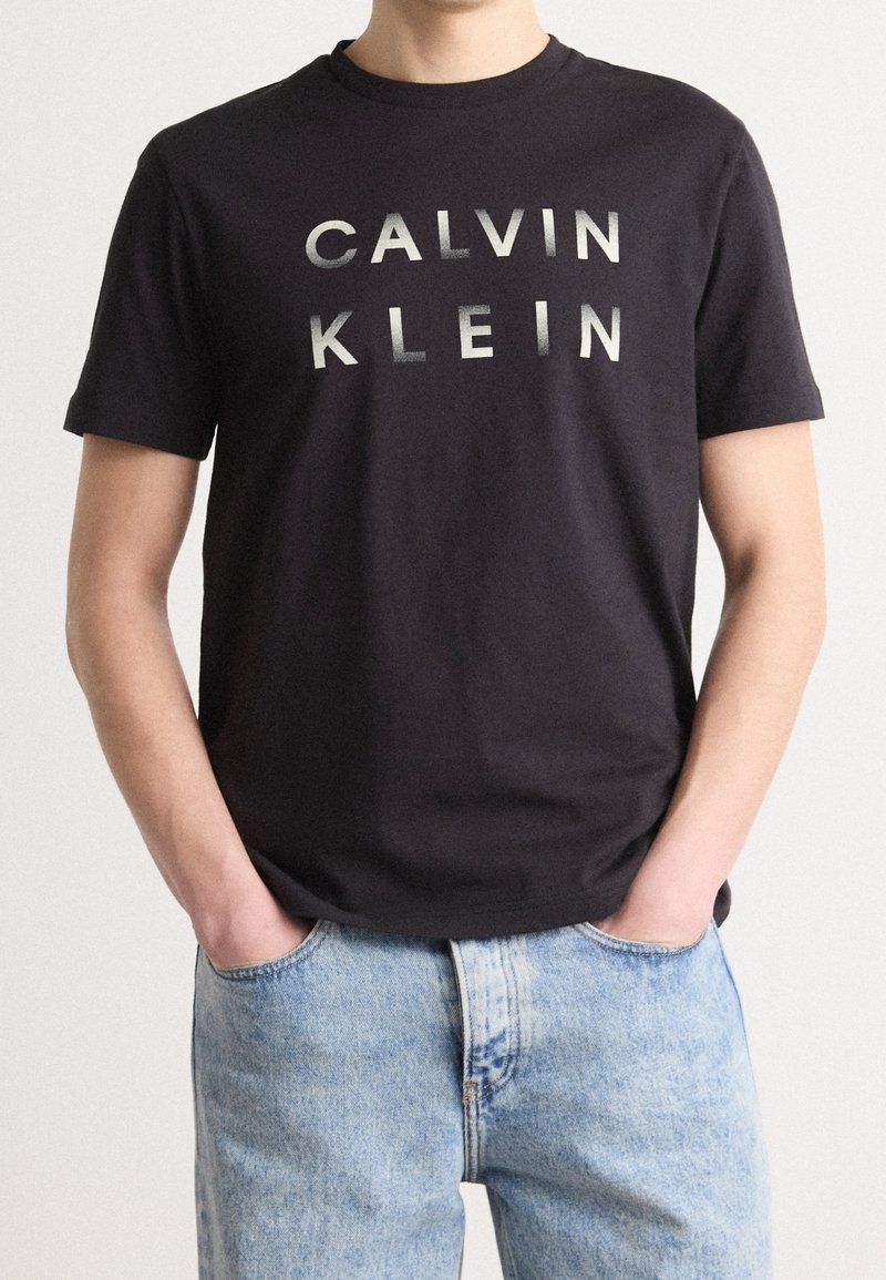 T-shirt en coton noir avec "CALVIN KLEIN" imprimé en texte argenté sur la poitrine. Manches courtes, col rond classique, associé à un jean bleu clair.