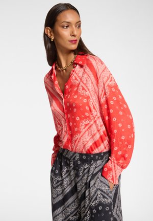 Blusa rossa a fantasia con disegni floreali e paisley, dotata di un colletto e chiusura con bottoni. Abbinata a pantaloni neri a fantasia.