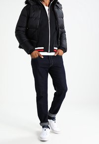 Black puffer jacket with a fur-lined hood over a dark zip-up sweatshirt, paired with rolled dark denim jeans and white sneakers.