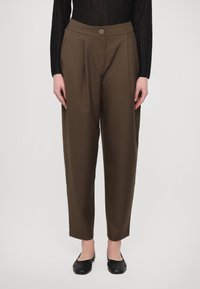 Kvinna klädd i högmidjade, avsmalnande bruna byxor med knappstängning, matchade med svarta ballerinaskor och en svart långärmad topp.