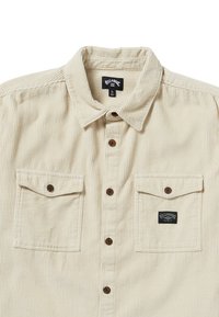 Beige kordfløjl knap-op skjorte med spids krave, to brystlommer og brune knapper. Har et sort logo patch på den venstre lomme.