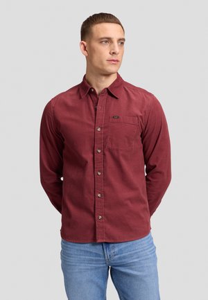 Uomo che indossa una camicia bordeaux a maniche lunghe con taschino sul petto e jeans azzurri, in piedi con le mani dietro la schiena.