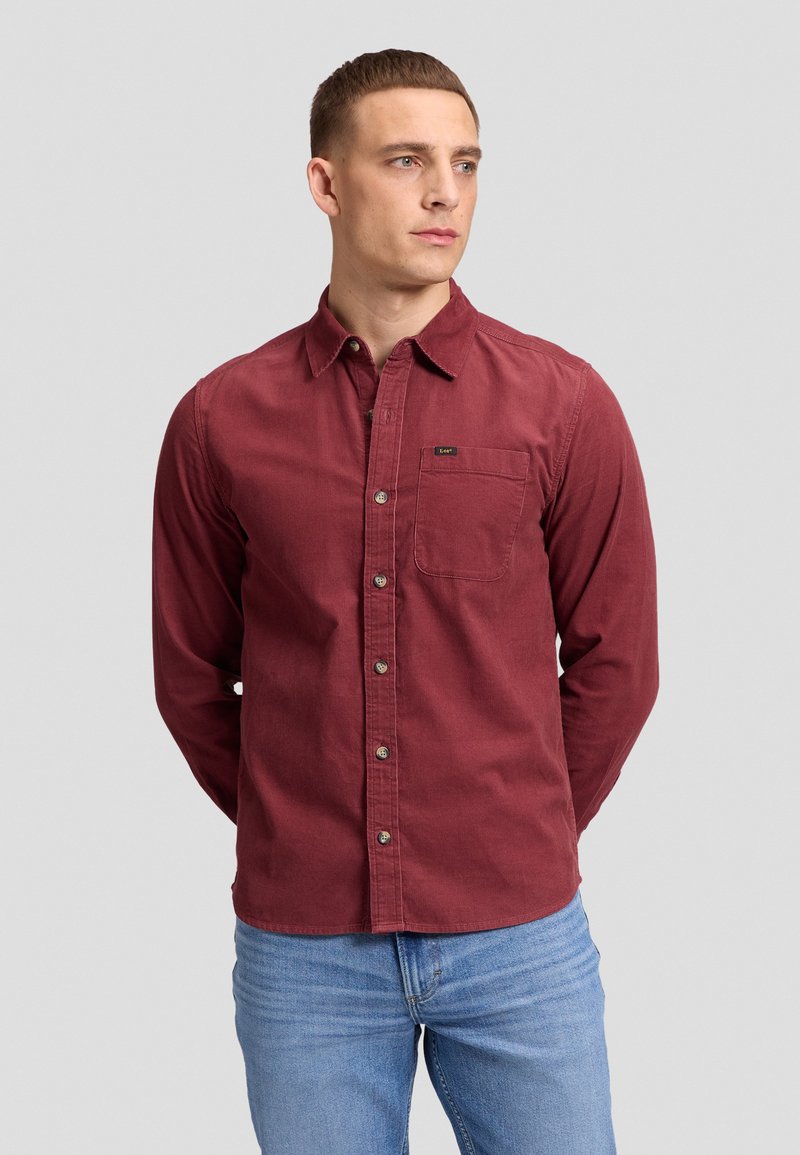 Homme portant une chemise bordeaux à manches longues avec une poche poitrine et un jean bleu clair, debout les mains derrière le dos.