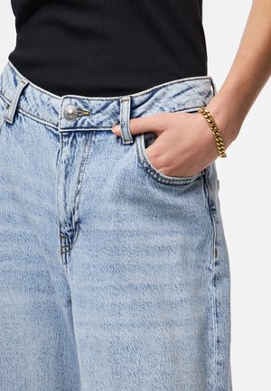 Hånd i forlomme på lyseblå denimjeans iført et guldarmbånd og en sort ribbet top.