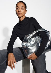 Metallisch silberne Handtasche mit glatter Textur und runden Griffen, gehalten von einer Person in einem schwarzen Langarmoberteil und grauen Hosen.