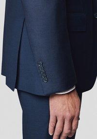 Granatowa marynarka o fakturowanej tkaninie, dopasowanym kroju i czterema czarnymi guzikami na mankietach, z widoczną białą koszulą pod spodem.