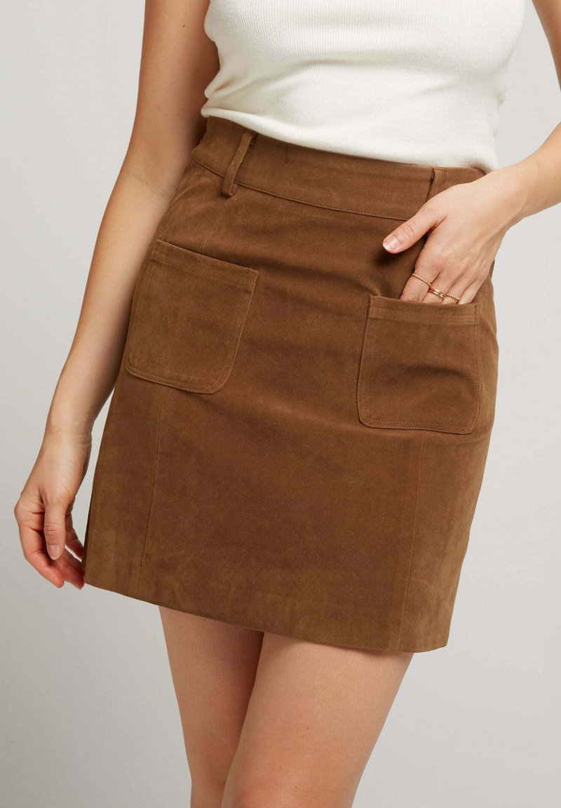 Jupe mini en daim marron avec deux poches plaquées à l'avant, une taille cintrée et une texture lisse. Longueur courte avec des détails de couture soignés.