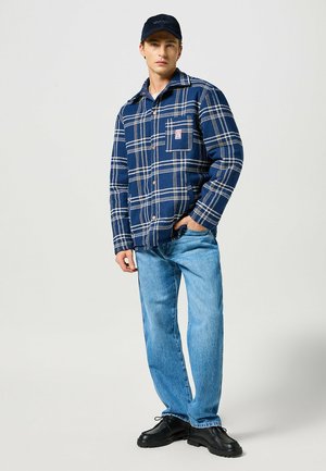 Giacca camicia a quadri blu navy con linee beige, bottoni e tasca sul petto, abbinata a jeans a gamba dritta blu chiaro e scarpe nere.