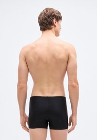 Μαύρα μαγιό σορτς που απεικονίζονται σε άτομο από πίσω, με απλό σχέδιο, μονόχρωμα και λεία υφή με εφαρμοστή κοπή.