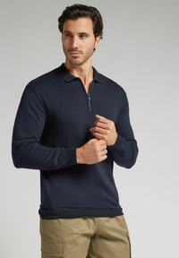 Granatowy sweter z długim rękawem z zamkiem przy kołnierzu, ściągaczem na mankietach oraz teksturowanym włóknem. Posiada dopasowany krój.