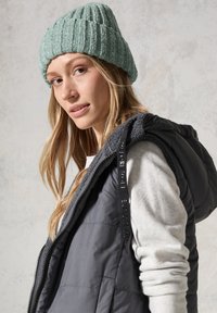 Grå, puffy väst med huva, bärs över en ljusgrå tröja. Modellen har på sig en texturerad mintgrön stickad mössa. Mjuka, avslappnade material.