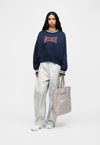 Γυναίκα που στέκεται, φοράει μπλε ναυτικό φούτερ Nike, ανοιχτό γκρι παντελόνι, λευκά αθλητικά παπούτσια, κρατώντας μπεζ τσάντα tote Nike σε απλό φόντο.