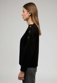Pull en maille noir avec un col rond, présentant des boutons sur les côtés, des détails de poignets côtelés et une coupe décontractée. Porté avec un jean gris.