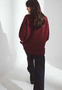 Burgunderfarbiges, übergroßes Sweatshirt mit geripptem Saum, aus weichem Material. Kombiniert mit dunklen, weiten Jeans, von hinten betrachtet, in sanftem Licht.