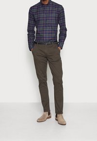 Camicia a quadri blu navy, verde e borgogna; pantaloni aderenti verde oliva; stivali alla caviglia in camoscio beige; texture liscia.