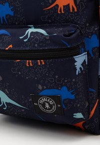 Primer plano de un bolsillo de mochila azul oscuro con cremallera negra, patrones coloridos de dinosaurios y un parche con el logo de Parkland Design & Manufacturing.