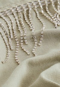 Plusieurs chaînes en strass argentées disposées en franges sur un tissu côtelé beige avec plis et texture visibles.