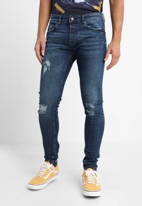 Vaqueros ajustados de mezclilla azul oscura, con detalles desgastados y un corte ceñido. Combinados con zapatillas amarillas, resaltando un estilo informal.