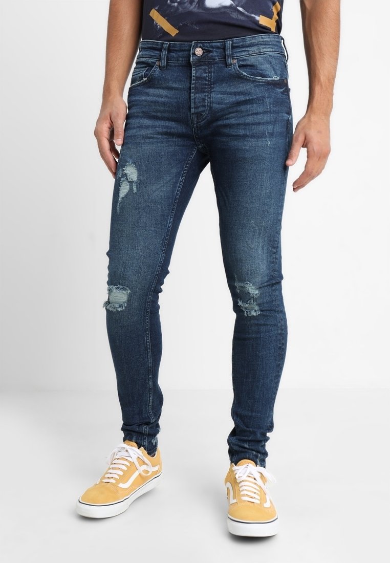 Vaqueros ajustados de mezclilla azul oscura, con detalles desgastados y un corte ceñido. Combinados con zapatillas amarillas, resaltando un estilo informal.