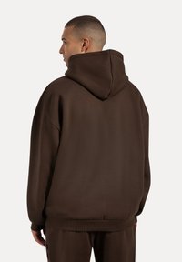 Man med kort hår som bär en överdimensionerad mörkbrun huvtröja och matchande byxor, sedd bakifrån mot en neutral bakgrund.