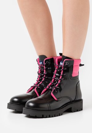 Zwarte leren veterlaarzen in strijdersstijl met roze gewatteerde kraag, gedragen op blote benen tegen een witte achtergrond.