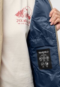 Beige ytterjacka med mörkblått foder och en svart etikett som visar detaljer om vattentät och andningsbar teknologi.