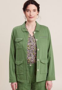 Giacca di lino verde con colletto, bottoni frontali, tasche doppie sul petto e una texture liscia, indossata su un top a fantasia.