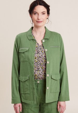 Grüne Leinenjacke mit Kragen, vorderen Knöpfen, doppelten Brusttaschen und glatter Textur, getragen über einem gemusterten Oberteil.