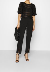 Svart genomskinlig topp med randiga volanger, kombinerad med figurnära svarta byxor med sidospalter. Modellen håller en svart kuvertväska och bär klackskor med remmar.