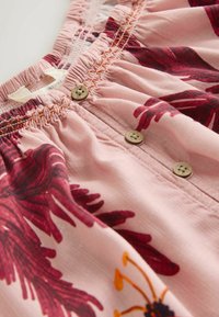 Roze stof met rood bloemenpatroon, elastische tailleband met plooien en drie bruine knopen in het midden.