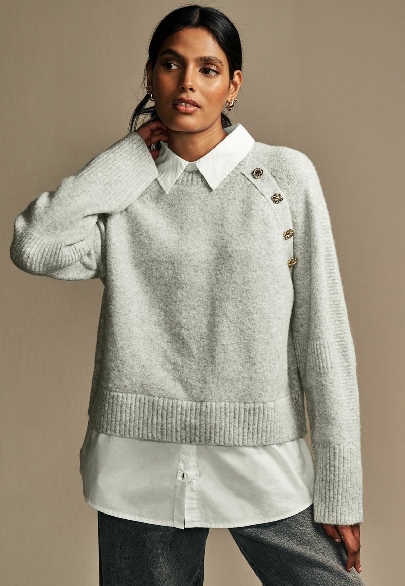 Lys grå strikket sweater med ribbet kant og manchetter, der har en hvid skjorte under. Guldknapper pryder skulderen.