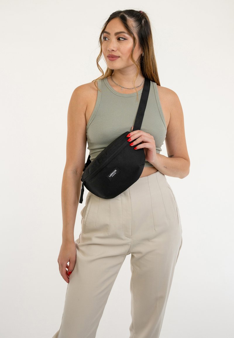 Čierna látková crossbody taška so zaobleným tvarom, zipsom a logom, nosená cez svetlošedý outfit so neutrálnymi tónmi.