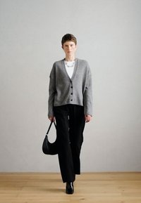 Cardigã cinza com botões, combinado com uma camisa branca e calças largas pretas. Uma bolsa preta e botas de tornozelo completam o visual.