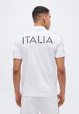 Homme portant un polo blanc et un short avec "ITALIA" inscrit en travers du haut du dos, face à un fond uni.