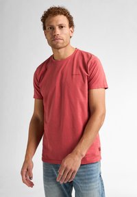 Camiseta de algodón de manga corta y cuello redondo en color coral, con detalles de costura ligeros, combinada con unos jeans azul desgastados. Marca minimalista en el pecho.