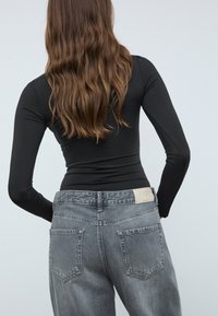 Čierny tričko s dlhým rukávom v kombinácii s vysokými vyblednutými sivými rifľami, ktoré majú béžovú logo náplasť na zadnej strane a dve zadné vrecká.