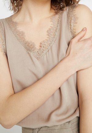 Frau trägt ein beige ärmelloses Oberteil mit Spitzenbesatz am Ausschnitt und an den Armlöchern und hält ihren linken Arm mit der rechten Hand.