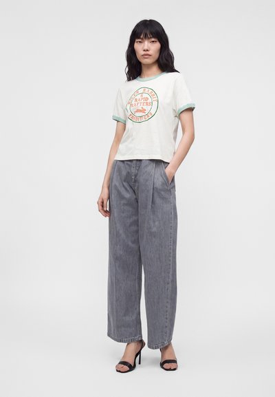 Femme portant un t-shirt graphique blanc avec un bord vert, un jean gris ample et des sandales noires à talons, debout avec une main dans la poche.