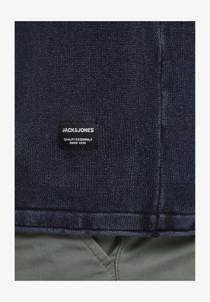 Tmavomodrý pletený sveter s rebrovaným lemom a manžetami, s čiernym štítkom s bielym textom "JACK&JONES" a "QUALITY ESSENTIALS SINCE 1990."