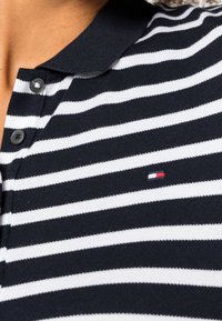 Personne portant un polo à rayures horizontales noires et blanches avec trois boutons noirs et un petit logo de marque rouge, blanc et bleu.